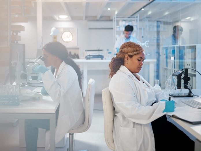 A pair of scientists work at their desks in a pharmaceutical research lab. One writes, while the other looks at a microscope.