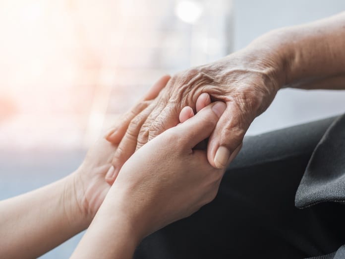 A close-up of two adult hands reaching up to gently support and grasp the wrinkled hand of an older person.