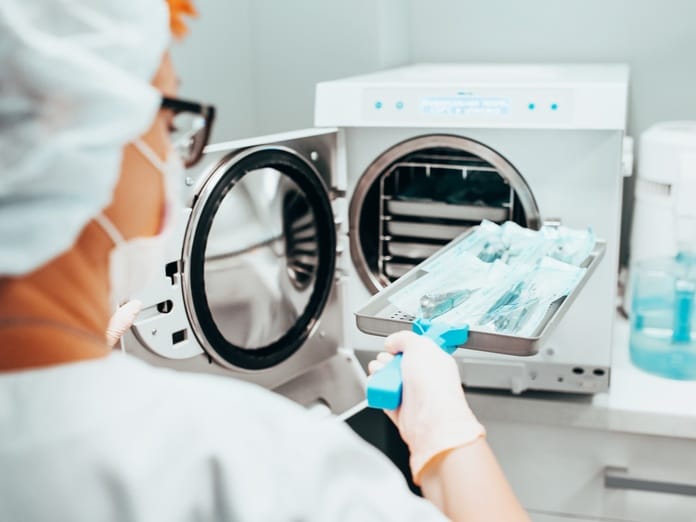 A medical professional placing a sterilizing tray full of tools into an autoclave. They're wearing a face mask and gloves.