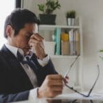 A man in a suit and tie sits at a desk, holding his glasses in one hand while pressing the bridge of his nose with the other.