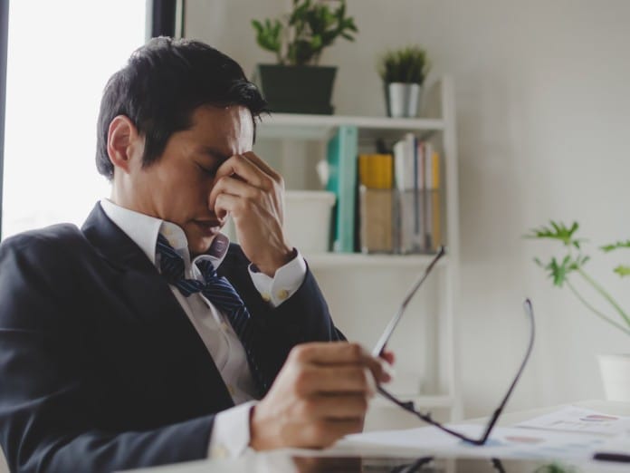 A man in a suit and tie sits at a desk, holding his glasses in one hand while pressing the bridge of his nose with the other.
