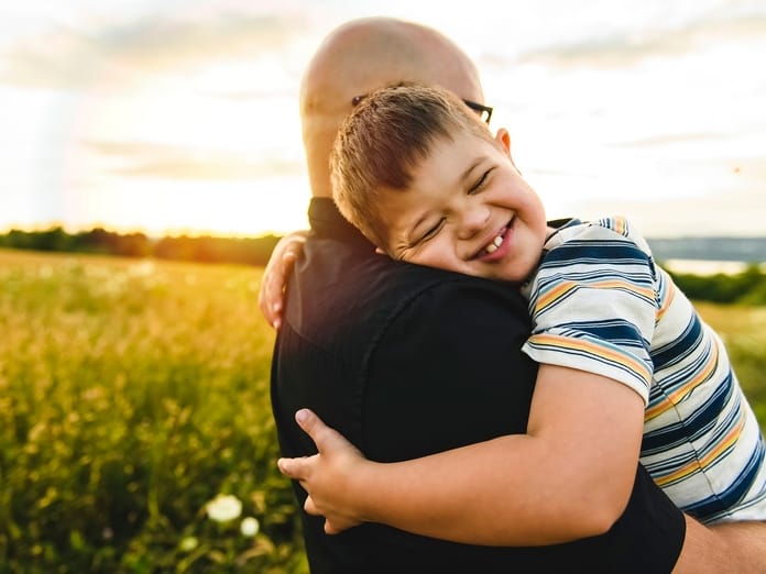 A young boy wearing a striped shirt smiles and closes his eyes while he hugs his dad tight. His dad is holding him.