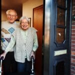 Two older adults with white hair and glasses stand at an opened door. One of them is holding on to a walker.