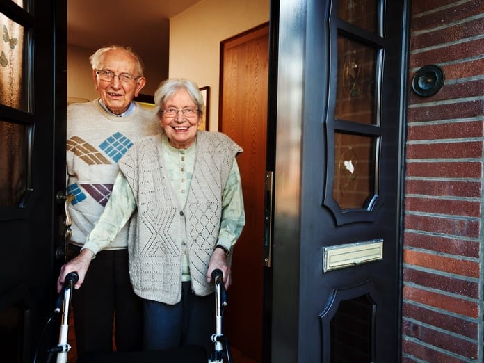 Two older adults with white hair and glasses stand at an opened door. One of them is holding on to a walker.