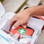 A hand grabbing a small red bag with a green circle and white plus sign on it from a large medical kit.