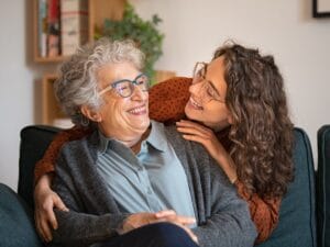 A woman leans over the back of a couch to wrap her arms around an older woman while they smile at each other.