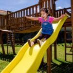 A young child excitedly waving their arms as they go down the bumps of a yellow playground slide.