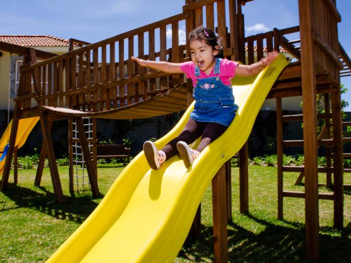 A young child excitedly waving their arms as they go down the bumps of a yellow playground slide.