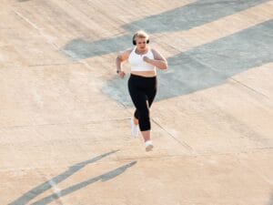 A woman in black leggings and a white sports bra and sneakers runs outside on a sunny day.