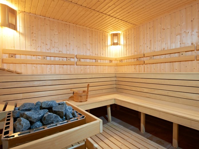 A sauna with wooden benches and walls, warm lighting, and a heater full of sauna rocks in the middle.