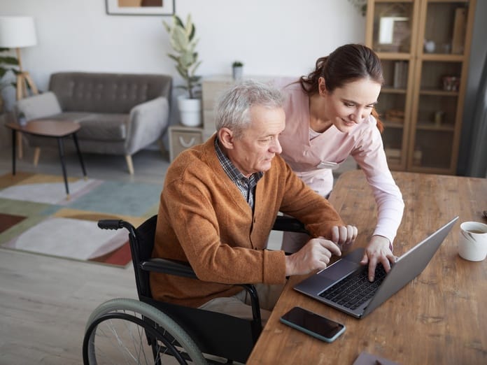 A senior man sitting in his wheelchair at the dining room table as a young female caregiver stands over his shoulder.