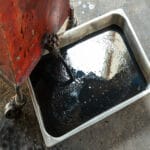 A stainless steel tray of motor oil laying on the ground. Someone is pouring more oil into the tray from a red can.