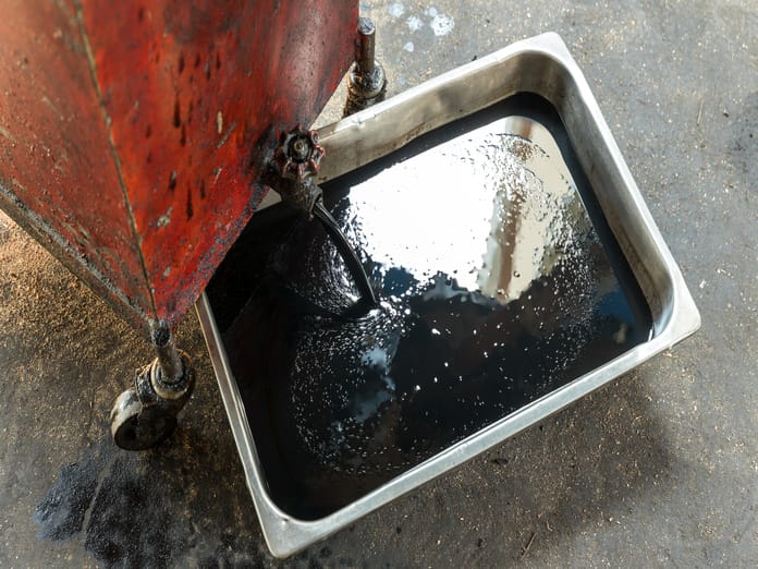 A stainless steel tray of motor oil laying on the ground. Someone is pouring more oil into the tray from a red can.