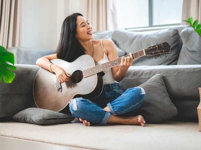 A smiling woman sits on the floor, wearing jeans and a white tank top, in front of a gray couch, playing an acoustic guitar.
