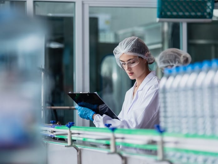 A female factory worker in a sterile uniform operates on the assembly line, inspecting bottles of water.
