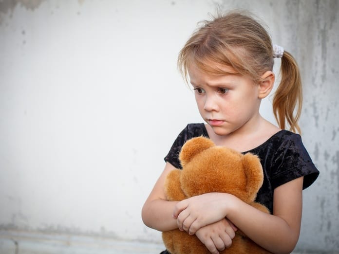 A young, anxious girl standing against a gray backdrop. She has pigtails and is clinging to a stuffed teddy bear.