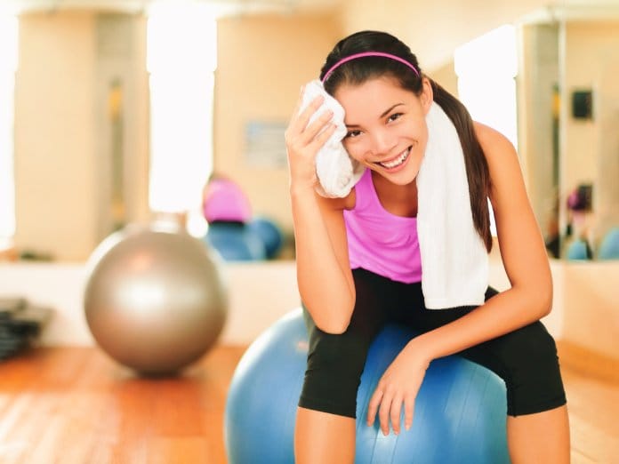 A smiling woman sitting on an exercise ball and using a thick white towel to wipe sweat off her face.