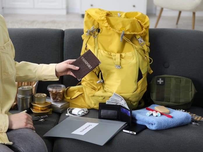 A person's hand holds a passport while sitting on a black sofa with a yellow backpack and emergency supplies on it.
