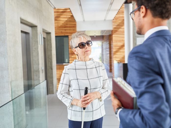 A visually impaired senior woman is wearing sunglasses and holding a cane. She is speaking to a man within a facility.
