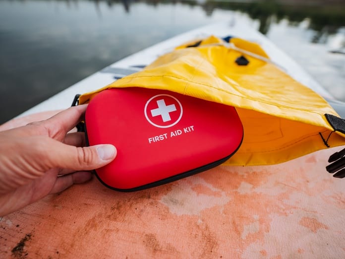 A person's hand holds a red first aid kit pouch next to a yellow bag sitting on top of a small boat.