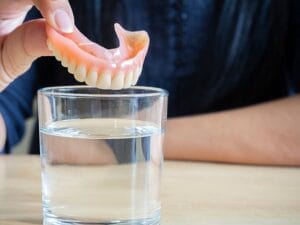 A close-up view shows a person's hand holding top denture teeth above a glass full of a clear liquid.