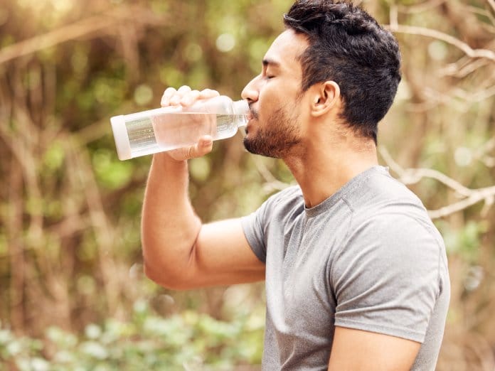 A man drinks from a bottle of clear water while standing in a wooded area. He is wearing a gray shirt.