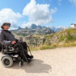 A man wearing a gray hat, sunglasses, and a black jacket sits in a black wheelchair on a gravel trail.