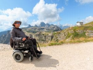 A man wearing a gray hat, sunglasses, and a black jacket sits in a black wheelchair on a gravel trail.