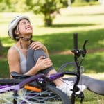 A woman sits on the ground next to a purple bicycle, clutching her knee. She has a painful look on her face.