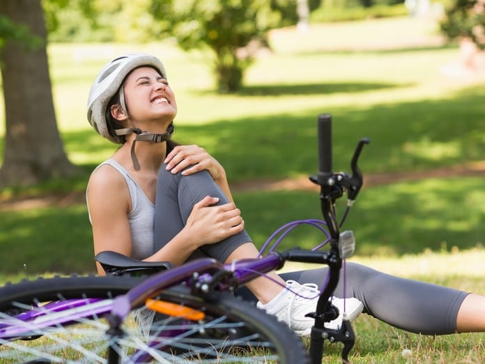 A woman sits on the ground next to a purple bicycle, clutching her knee. She has a painful look on her face.