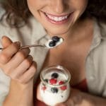 A close-up of a woman smiling while holding a spoon with yogurt and berries. She has the yogurt cup in her other hand.