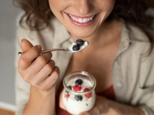 A close-up of a woman smiling while holding a spoon with yogurt and berries. She has the yogurt cup in her other hand.