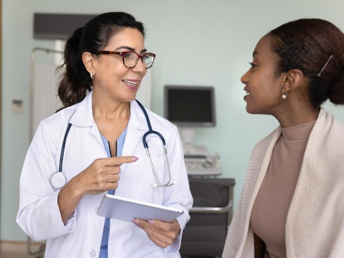 A mature woman doctor is speaking to her patient, a young woman, in the doctor's office. They're smiling at each other.