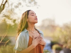 A woman is standing in a field and is holding her hands on her chest to use calming breathing practices.