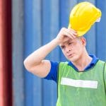 A man holds a hard hat above his head while wiping his forehead with his hand. He wears a blue shirt and a yellow vest.