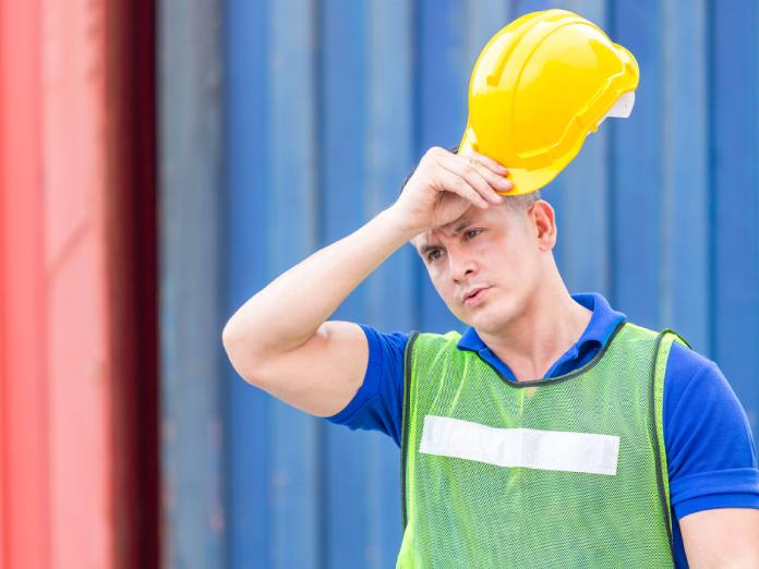 A man holds a hard hat above his head while wiping his forehead with his hand. He wears a blue shirt and a yellow vest.