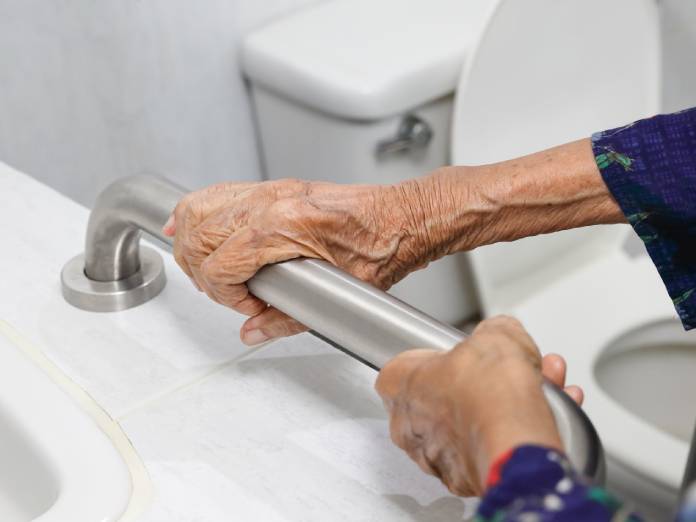 An older adult holding a stainless steel grab bar mounted next to a bathroom sink with a toilet in the background.