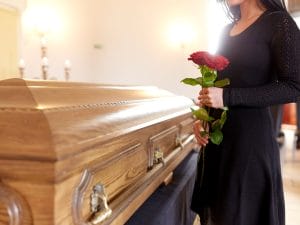 A young woman with long hair is wearing a black dress and holding two roses. She's standing next to a closed brown casket.