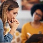 A nervous therapy client sits with her hands folded by her mouth. The therapist is behind her talking with a clipboard.