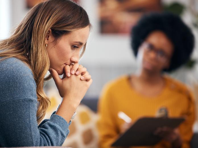 A nervous therapy client sits with her hands folded by her mouth. The therapist is behind her talking with a clipboard.