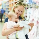 A smiling blonde woman holds her baby to her side as she examines a tiny white onesie in a store.