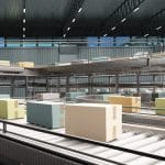 A large warehouse contains a conveyor belt with blue, yellow, orange, and brown cardboard boxes on it.