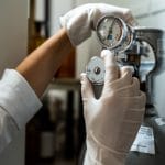 A close-up view shows a person wearing white latex gloves adjusting a stainless steel regulator on a gas canister.