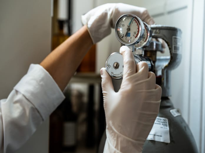 A close-up view shows a person wearing white latex gloves adjusting a stainless steel regulator on a gas canister.