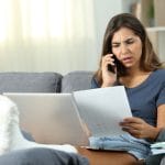 A young female patient sits on her couch with her ankle wrapped and elevated. She's on the phone arguing about a paper bill.