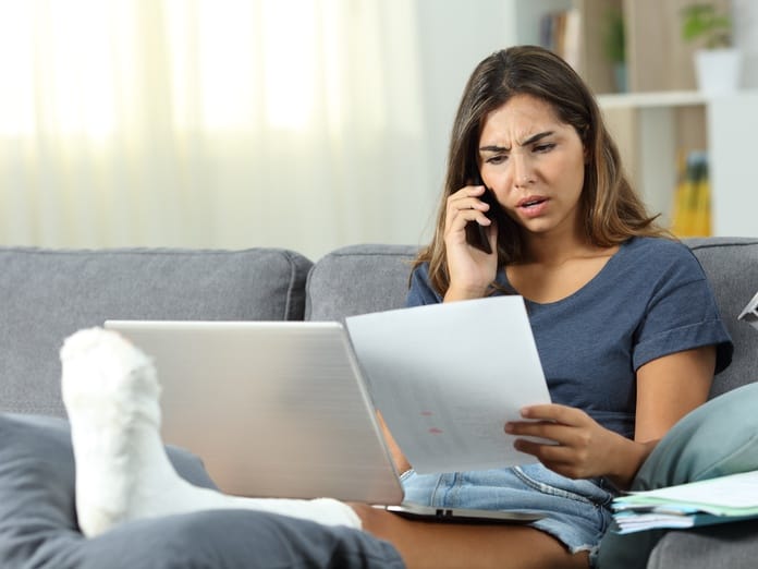 A young female patient sits on her couch with her ankle wrapped and elevated. She's on the phone arguing about a paper bill.