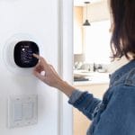 A female homeowner stands at her smart thermostat to adjust the system settings. There's a double-switch faceplate under it.