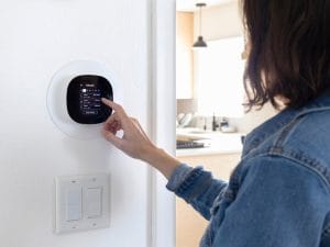 A female homeowner stands at her smart thermostat to adjust the system settings. There's a double-switch faceplate under it.