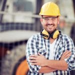A young construction worker wearing a checkered shirt and a hard hat crosses his arm and smiles while on a job site.