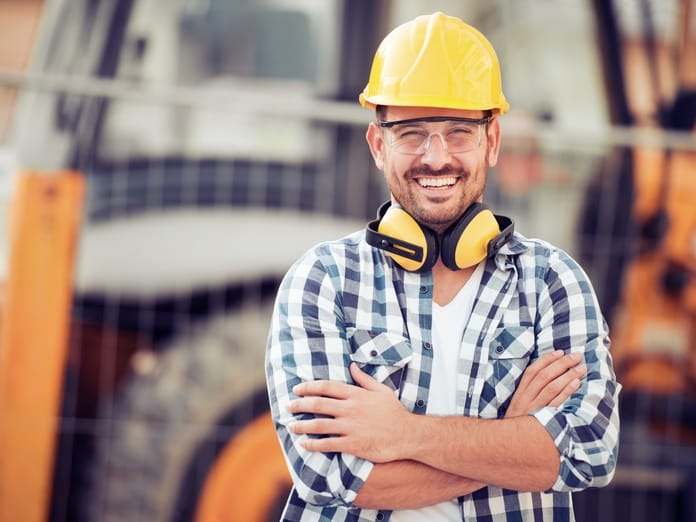 A young construction worker wearing a checkered shirt and a hard hat crosses his arm and smiles while on a job site.